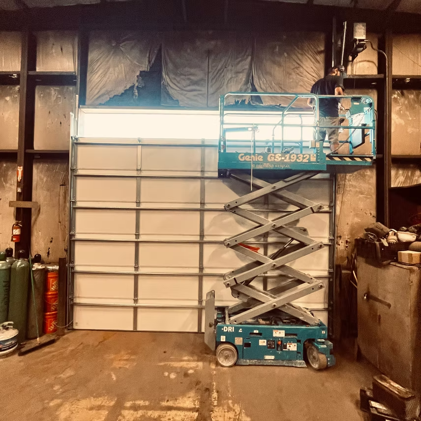 A person stands on a blue scissor lift working on a large white industrial garage door inside a workshop.