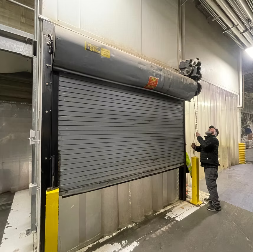 A person in a warehouse pulls a chain to operate a large, gray, metal industrial roll-up door.