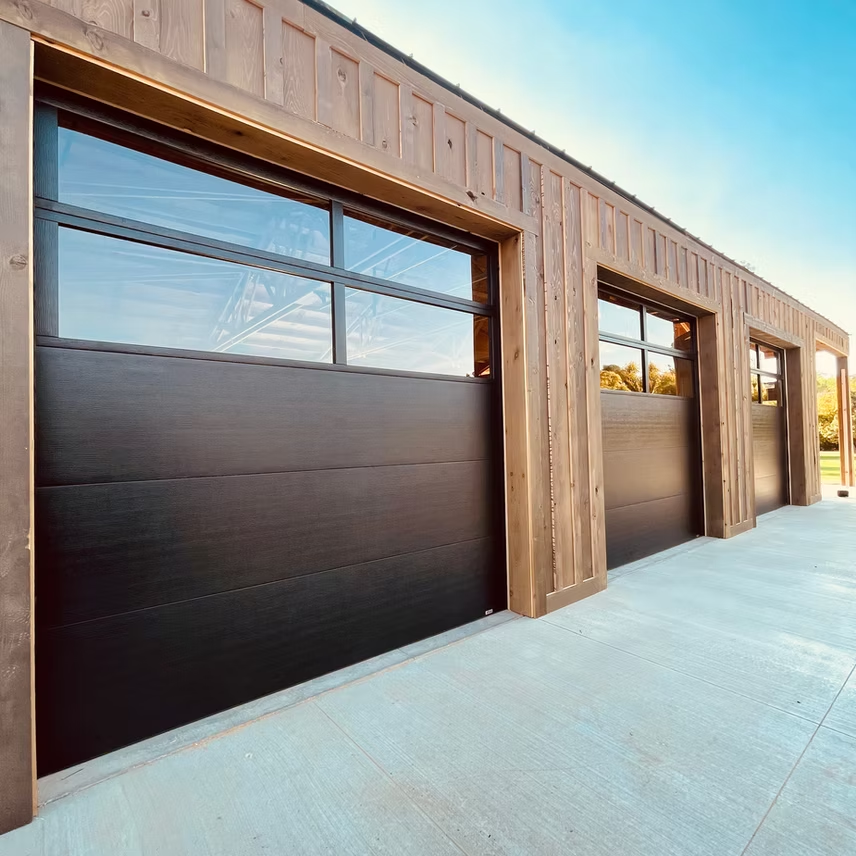 A row of modern, dark metal garage doors with top glass panels on a building with vertical wooden exterior siding.