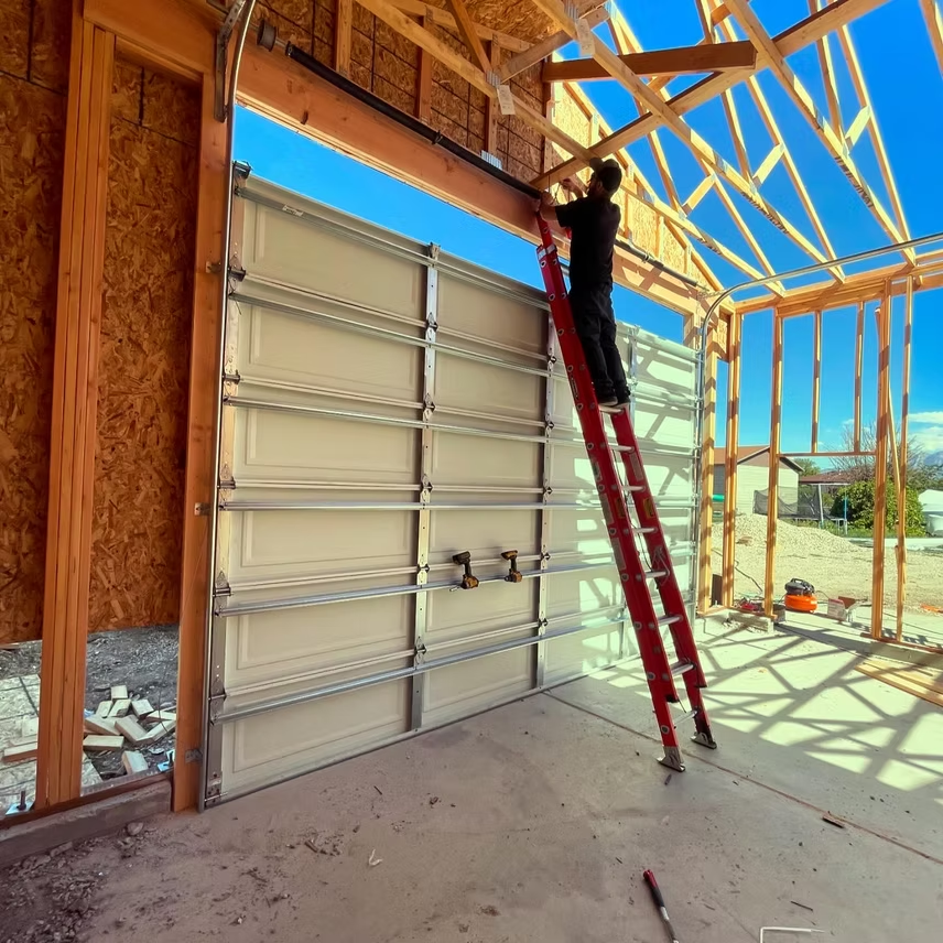 A person on a red ladder installs an overhead garage door in an unfinished wooden building frame under a clear blue sky.