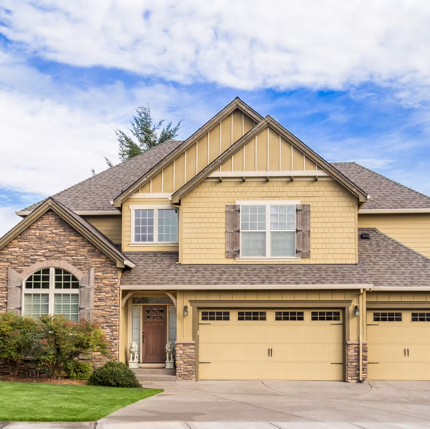 Two-story suburban home with tan siding, stone accents, a multi-car garage, and a peaked roof under a blue, cloudy sky.