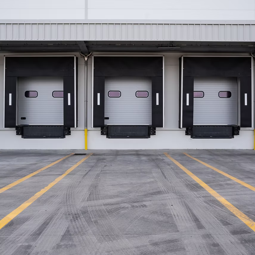 Three industrial loading dock bays with white closed doors and black seals on a concrete lot with yellow parking lines.