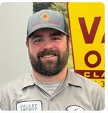 A person with a beard and a gray cap bearing a logo, smiling in front of a yellow and red Valley Energy sign.