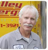 A person in a gray work shirt stands in front of a yellow truck with red lettering.