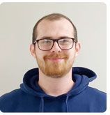 A person with a beard and glasses smiles at the camera, wearing a navy blue hoodie against a light gray background.
