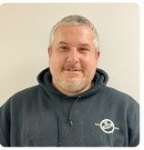 A smiling person wears a dark grey hooded sweatshirt with a small, circular white logo on the chest against a beige wall.