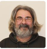 A smiling person with long gray hair, a full beard, and glasses, wearing a dark hooded sweatshirt against a plain background.