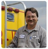 A person in a gray work shirt stands smiling in front of a yellow utility truck with a ladder.
