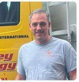 A man in a grey t-shirt smiles in front of a yellow International brand truck.
