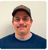 A person with a mustache and glasses wears a blue sweatshirt and a grey baseball cap against a plain white background.