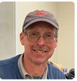 A smiling person with glasses wearing a grey cap and a blue sweater, shown from the chest up.