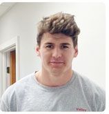 A person with short, wavy brown hair wears a gray crew-neck shirt against a white indoor background.