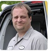 A person in a light grey work uniform smiling slightly, standing in front of a service vehicle.