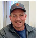 A smiling person wears a grey baseball cap with a red and gold logo and a dark collared jacket over a blue shirt.