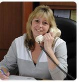 A person with blonde hair smiles while holding a telephone receiver to their ear and writing on a paper at a desk.