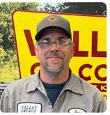 A person in a gray Valley Energy uniform and cap stands in front of a yellow sign.