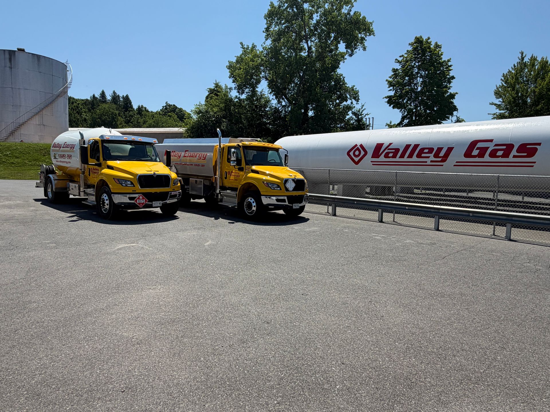 A yellow semi-truck with a stainless steel tanker trailer parked beside large industrial storage tanks outdoors.