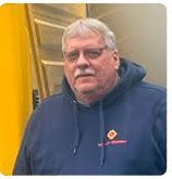 A person with gray hair and glasses wearing a blue hooded sweatshirt with a small logo, standing indoors.