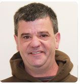 A person with short, dark hair wears a brown hoodie, smiling slightly against a plain white background.