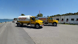 A yellow semi-truck with a stainless steel tanker trailer parked beside large industrial storage tanks outdoors.