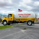 A yellow tanker truck is parked in front of several large, cylindrical storage tanks under a cloudy sky.