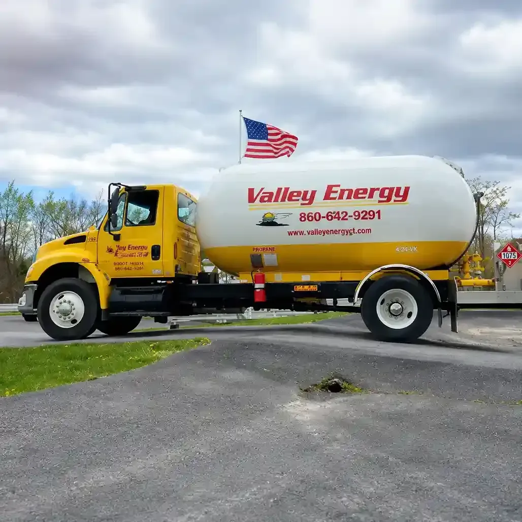 A yellow tanker truck is parked in front of several large, cylindrical storage tanks under a cloudy sky.
