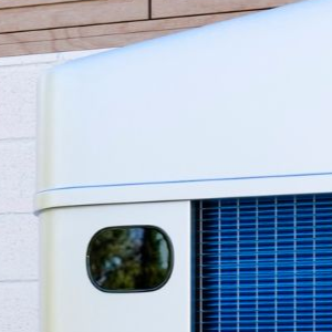 A modern air source heat pump unit standing on a concrete base against a white brick wall outside a house.