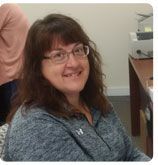 A person with brown hair and glasses smiles while wearing a grey quarter-zip pullover in an office setting.