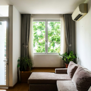 A room featuring a brown sofa, a large window with gray curtains overlooking green trees, and an air conditioning unit.