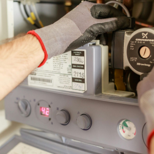 A technician wearing work gloves repairs a wall-mounted gas boiler, adjusting a circular pump component.