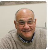 A smiling individual with glasses wearing a beige sweater against a neutral background.
