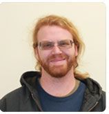 A smiling person with reddish-brown hair and a beard, wearing glasses and a dark hooded jacket against a neutral background.