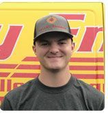 A smiling person wearing a gray cap and dark shirt, positioned in front of a yellow van with red lettering.