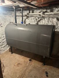 A metal residential heating oil tank sits in a basement against a stone wall, showing signs of staining and oil leakage.