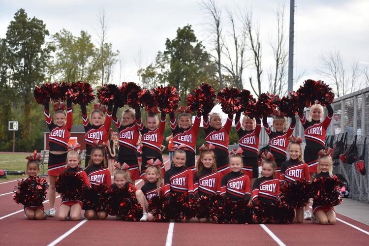 Cheerleading team posing on a track in red uniforms with pom-poms, trees and bleachers behind them