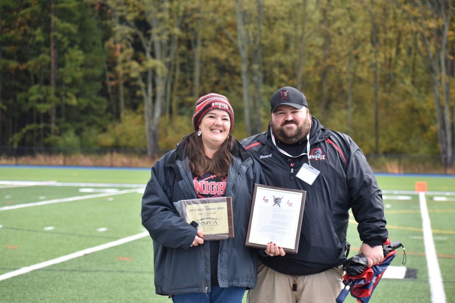 Two people holding plaques on a sports field, smiling in front of a wooded background.