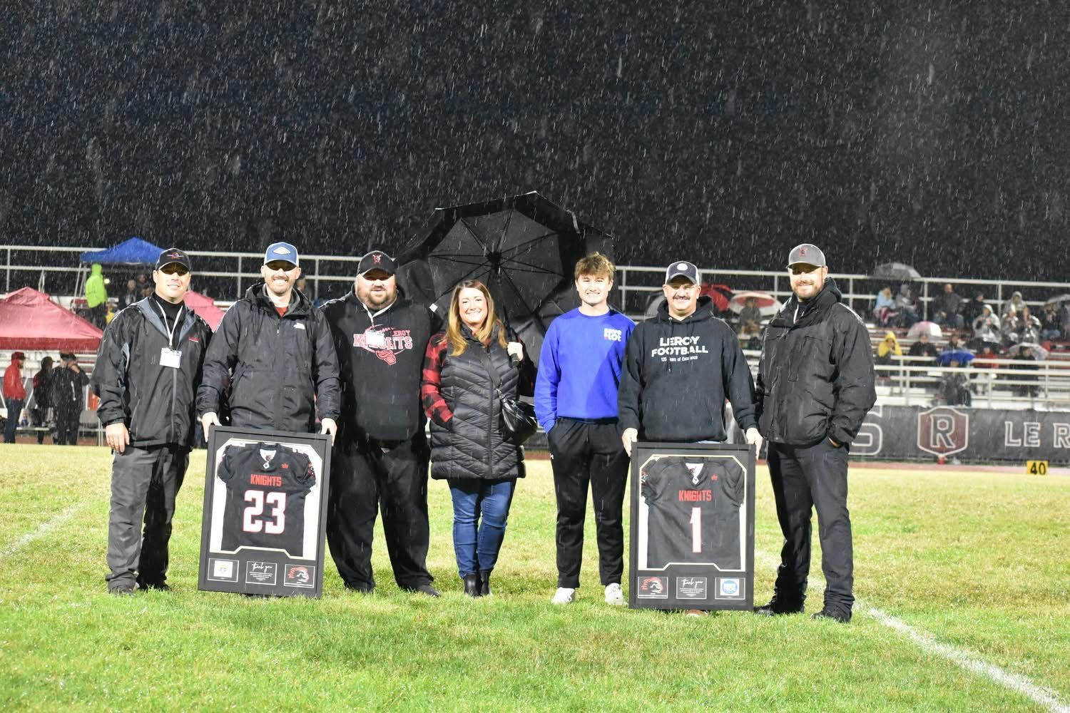 People standing on a rainy football field holding framed jerseys during a pregame ceremony