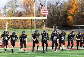 Youth football players in black uniforms lined up on a field beneath a goalpost and American flag.