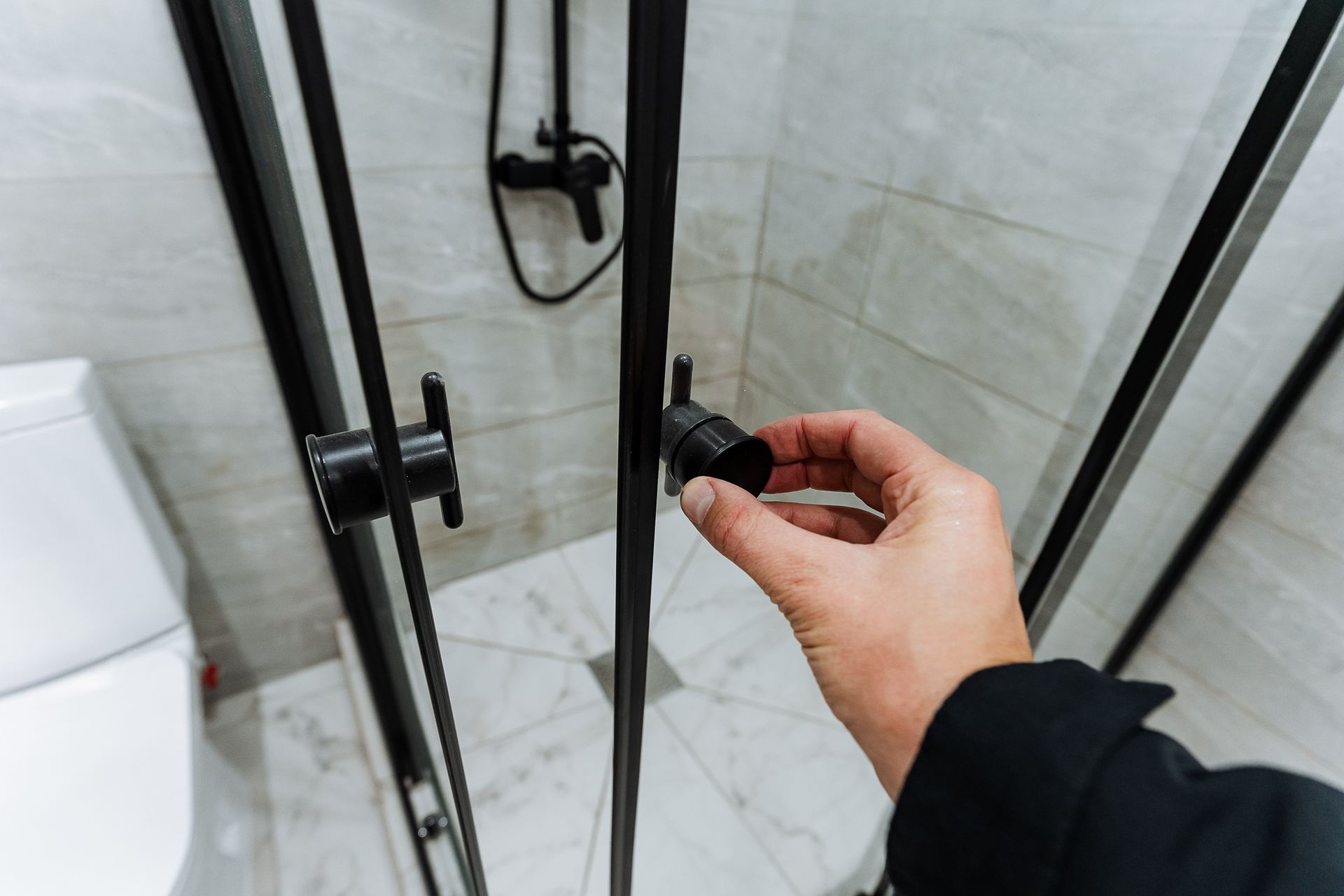 A hand holds the latch of a shower door inside a bathroom.