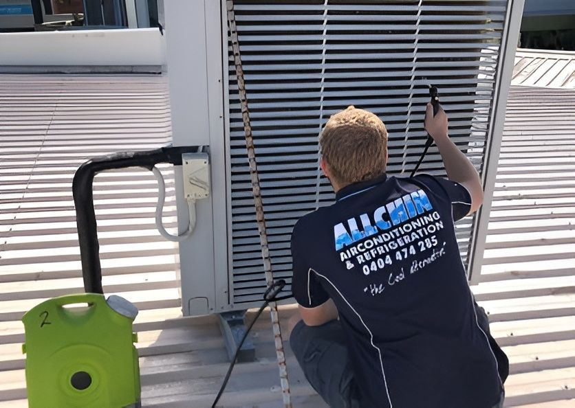 A Man In A Work Shirt Cleaning An Air Conditioning Unit On A Rooftop, Using A Hose And Spray Nozzle — Allchin Airconditioning & Refrigeration Electrical in Caloundra, QLD