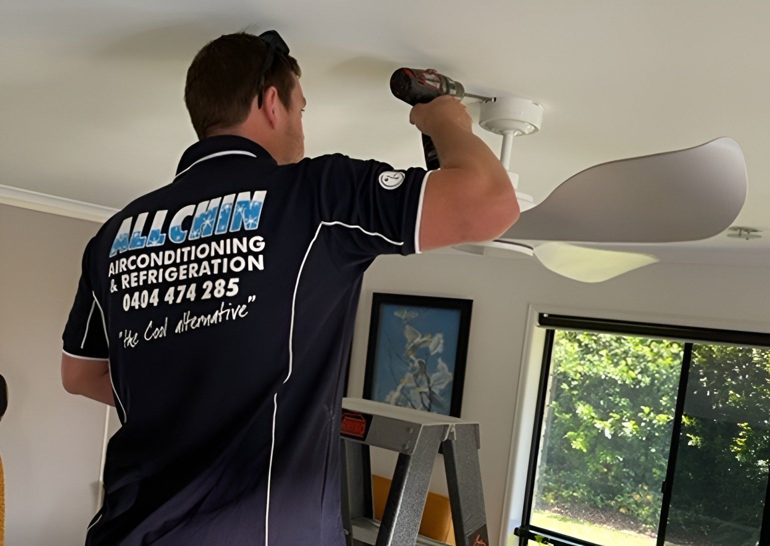 A Man In A Blue Uniform, Using A Drill To Install A White Ceiling Fan — Allchin Airconditioning & Refrigeration Electrical in Caloundra, QLD