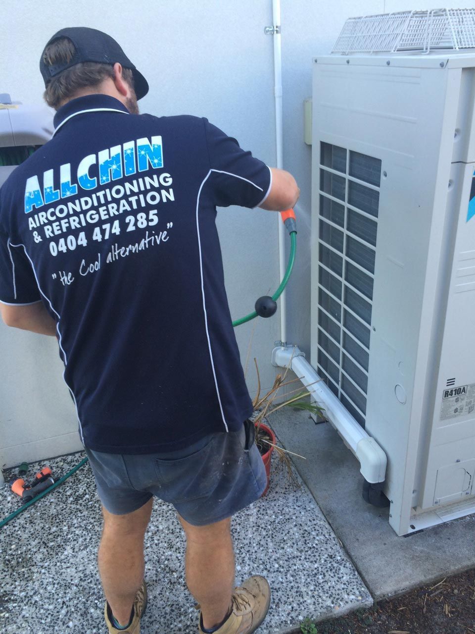 Man In Blue Work Shirt, Washing An Outdoor Air Conditioning Unit With A Hose — Allchin Airconditioning & Refrigeration Electrical in Caloundra, QLD