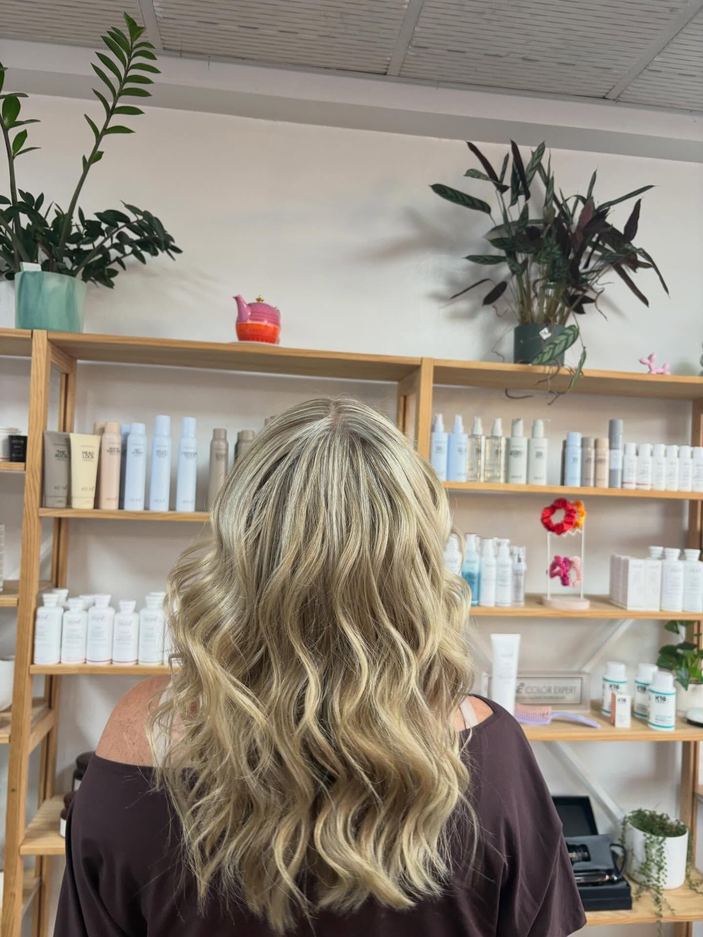 Person with wavy blonde hair seen from behind in a salon with product shelves and plants