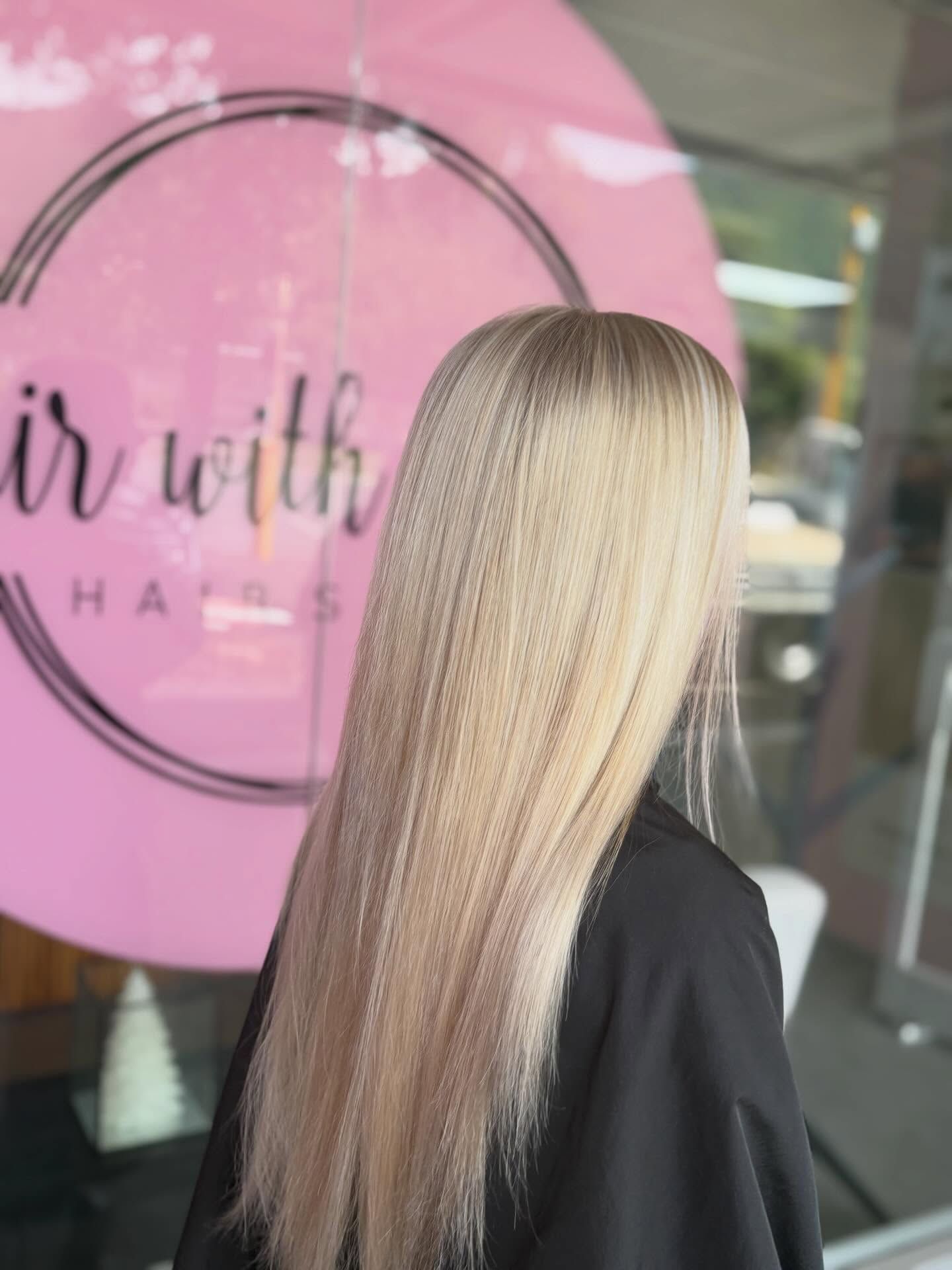 Long blonde hair with subtle highlights, shown from behind in a salon with a pink sign.
