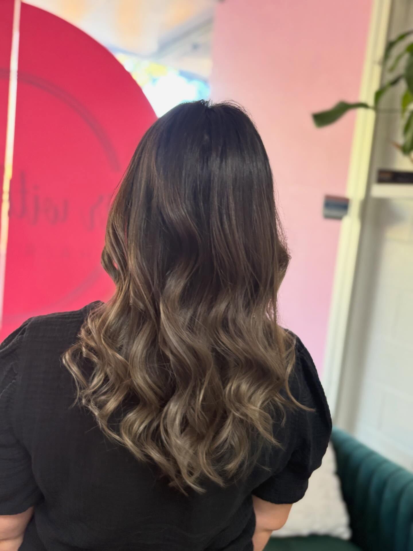 Woman with wavy brown hair from behind, in a salon with a pink wall and mirror.