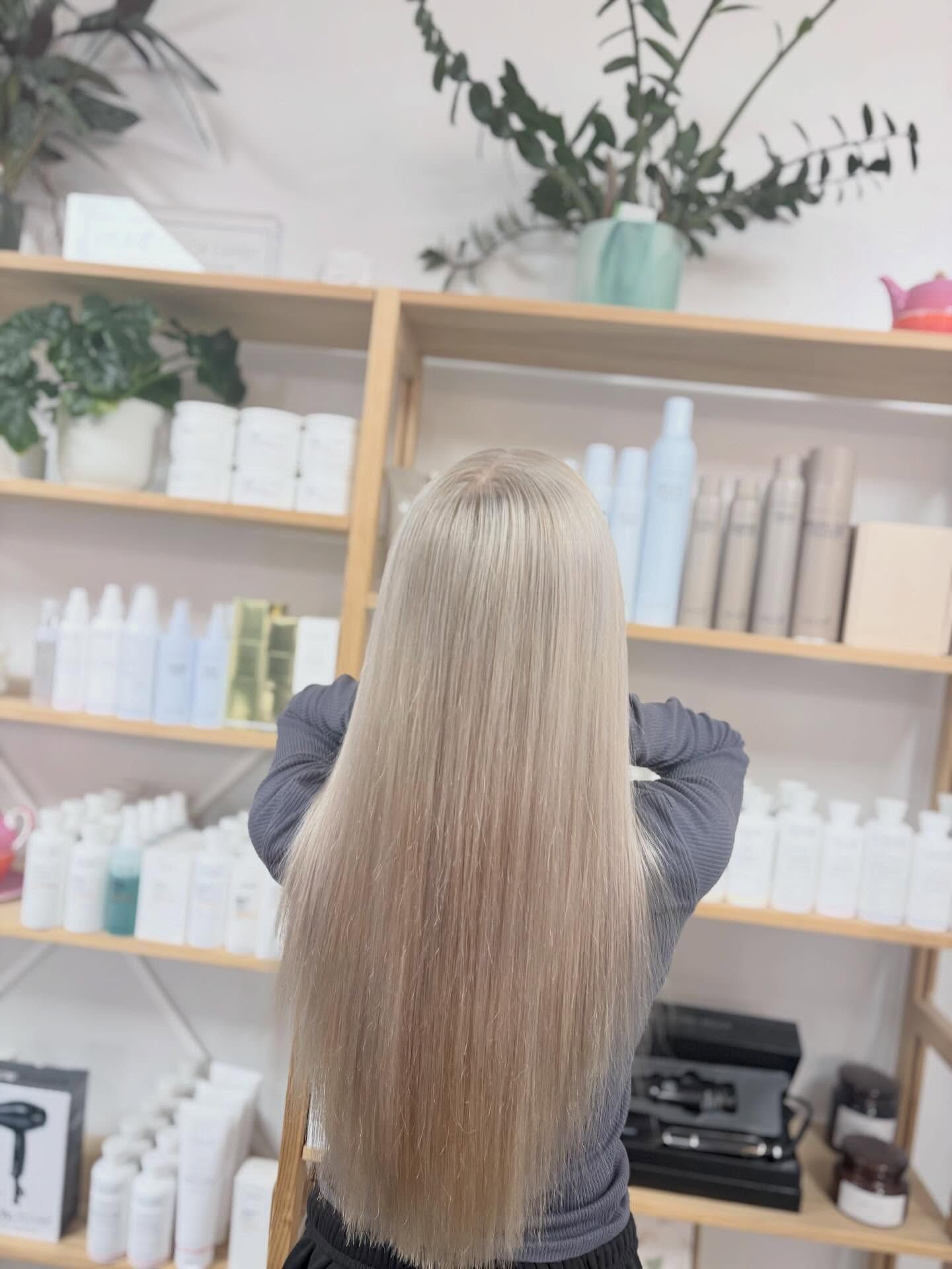 Person with long blonde hair standing in a salon, facing shelves of hair products.