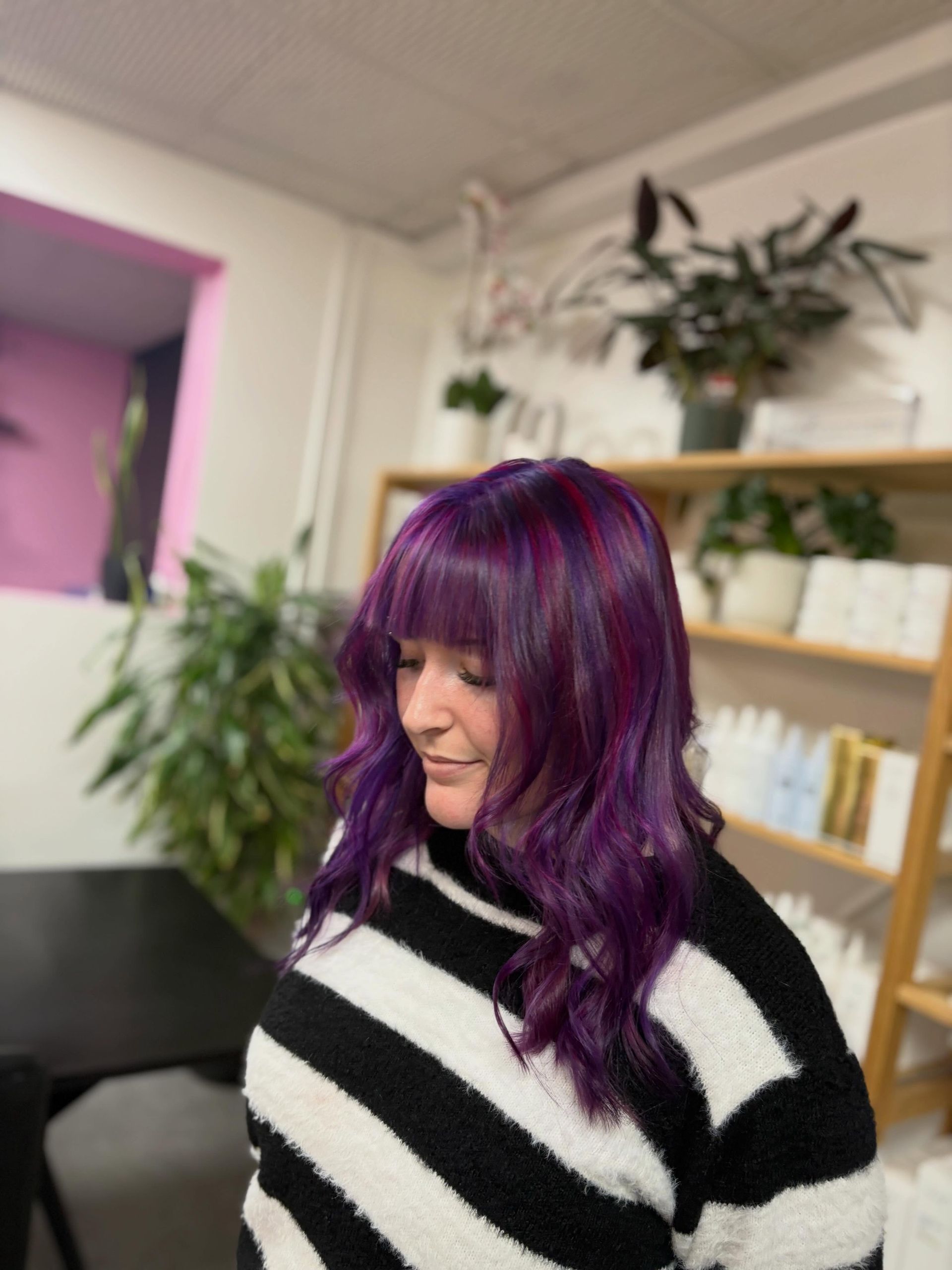 Person with purple hair in a black-and-white striped sweater in a salon, looking down