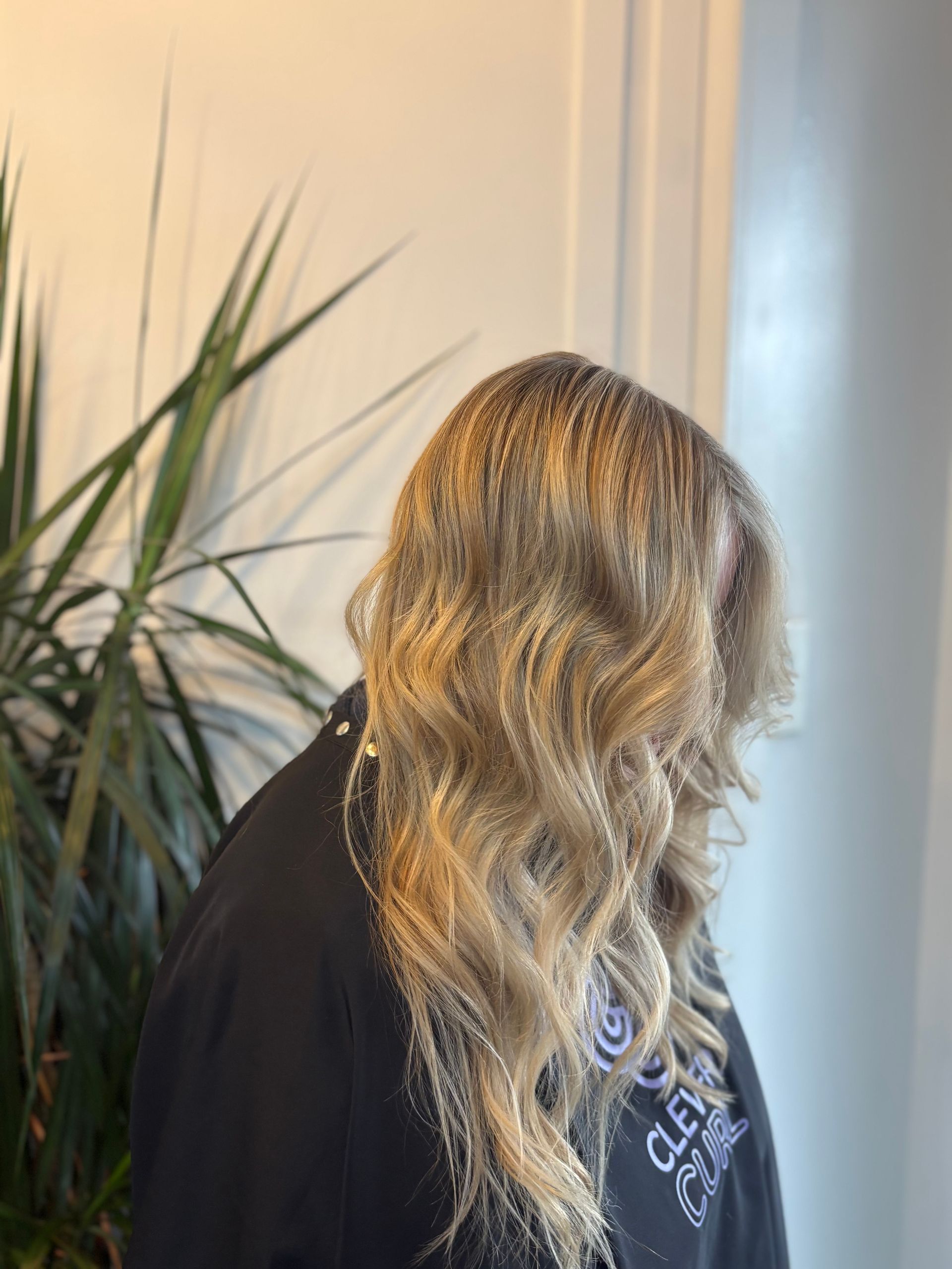 Person with wavy blonde hair in a black top, turned away indoors beside a leafy plant.