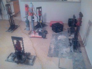 Multiple hydraulic jacks and a diamond core drill are positioned on a concrete floor inside a room.