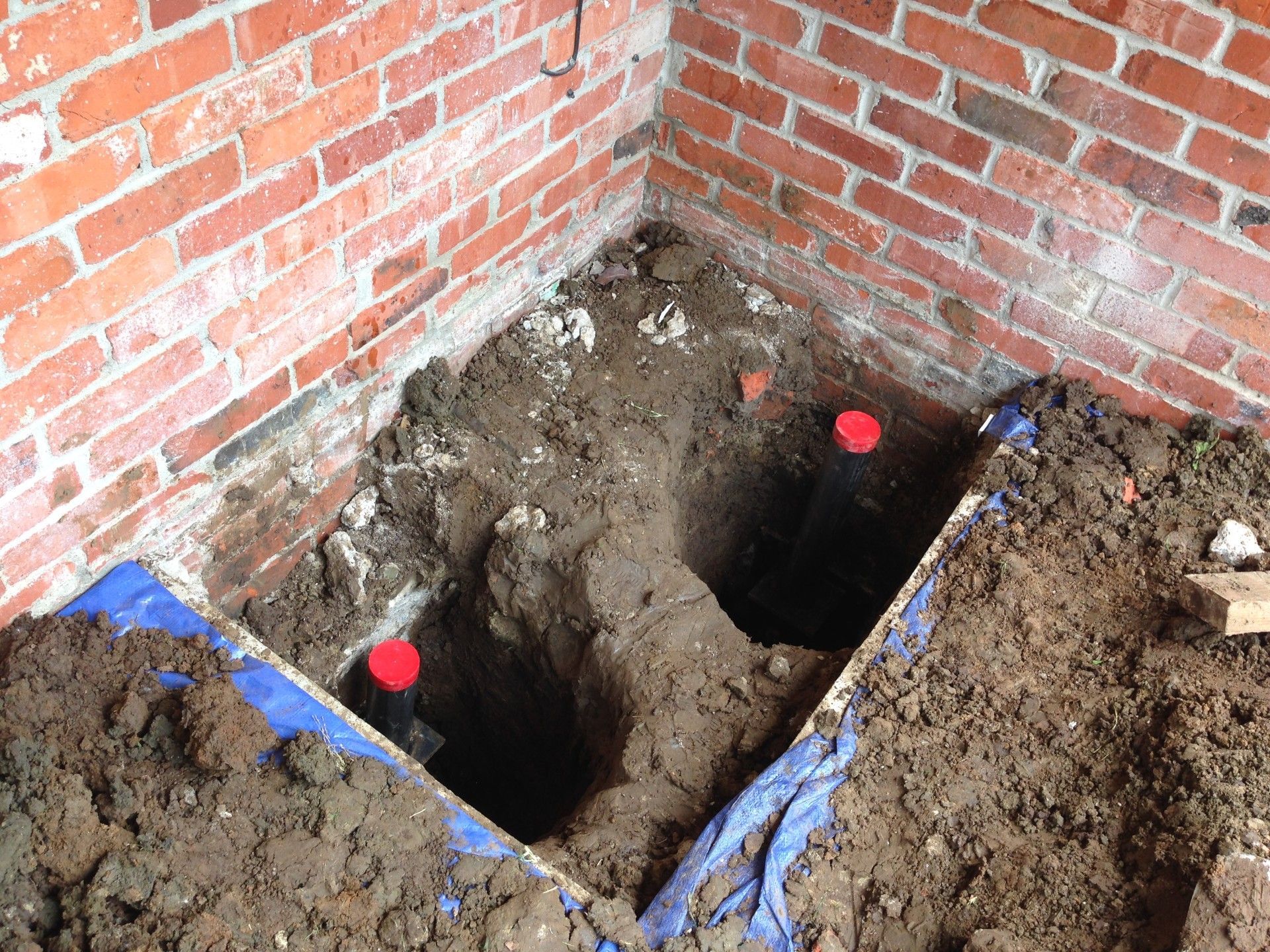Two vertical pipes with red caps stand in a dirt excavation area next to a red brick corner wall.